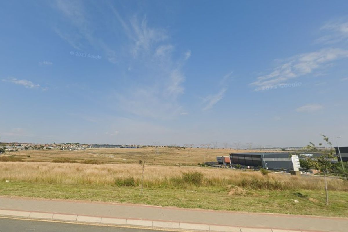 A view of an undeveloped commercial land plot from the adjacent street, featuring a paved sidewalk and surrounding industrial development.