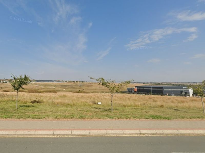 A view of a vacant commercial land plot from the adjacent street, featuring a paved sidewalk and open grass terrain.
