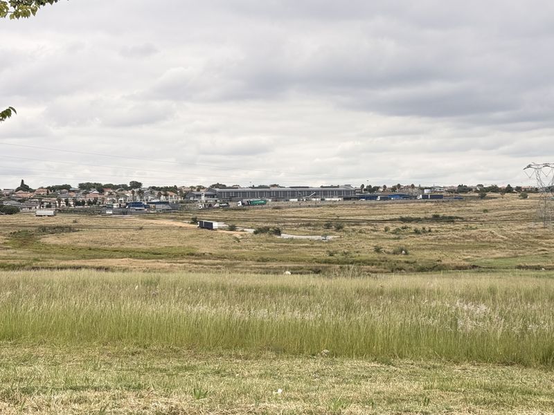 A wide-angle view of an expansive vacant commercial land plot featuring natural grassland and distant industrial infrastructure under a cloudy sky.