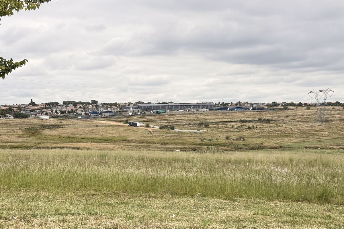 A wide-angle view of an expansive vacant commercial land plot featuring natural grassland and distant industrial infrastructure under a cloudy sky.