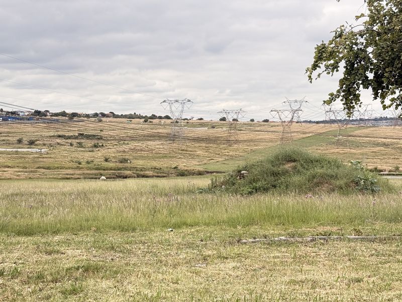 A wide view of open vacant land featuring rolling grass fields and prominent high-voltage electricity pylons crossing the landscape.