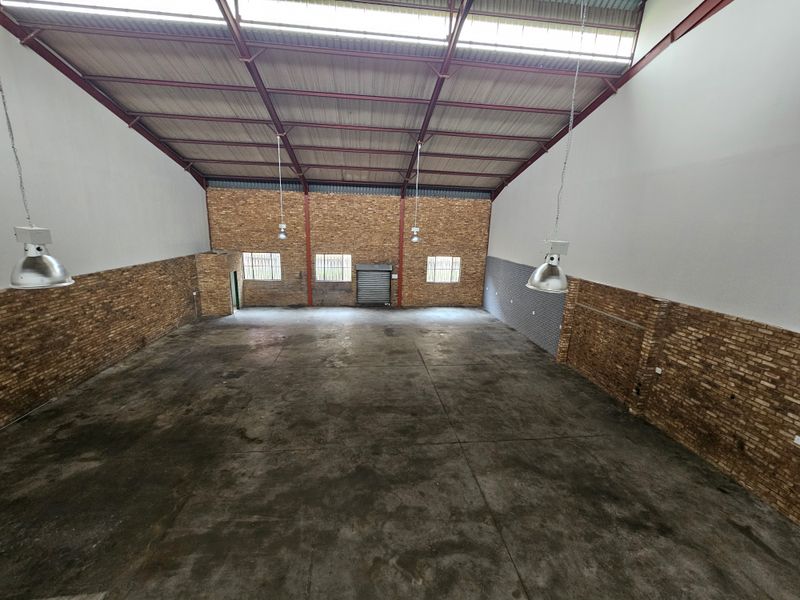 Spacious industrial warehouse unit featuring exposed steel roof trusses, brick walls, concrete flooring, and high-bay pendant lighting with a secure roller shutter door.