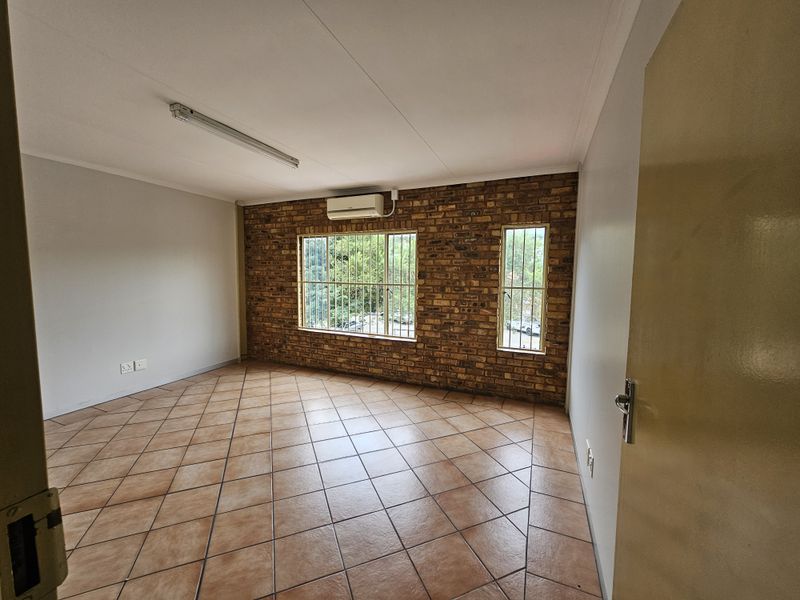 A clean, empty office space featuring an exposed brick wall, tiled flooring, security bars on windows, and a wall-mounted air conditioning unit.