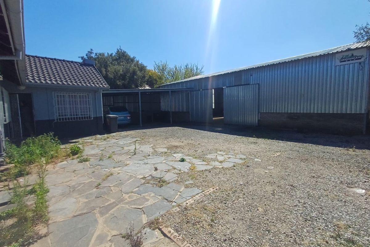 Rear exterior view of the industrial property featuring a large corrugated metal warehouse, a covered carport, and a combination of stone paving and gravel yard.