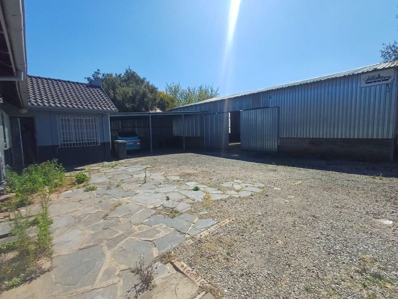 Rear exterior view of the industrial property featuring a large corrugated metal warehouse, a covered carport, and a combination of stone paving and gravel yard.