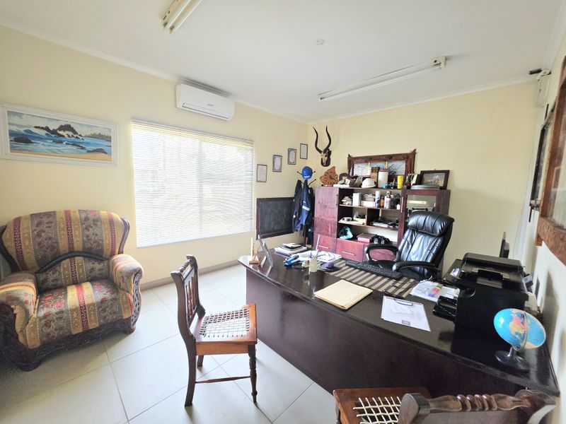 A functional office space featuring tiled flooring, a wall-mounted air conditioning unit, and large windows with venetian blinds for natural light.