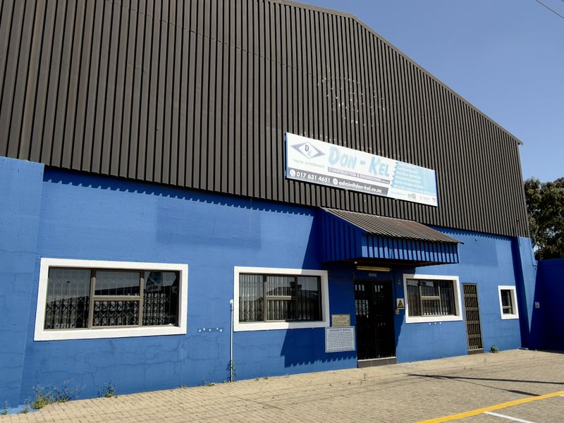 Front facade of a commercial industrial building featuring blue painted masonry, metal cladding, security window bars, and a paved parking area.