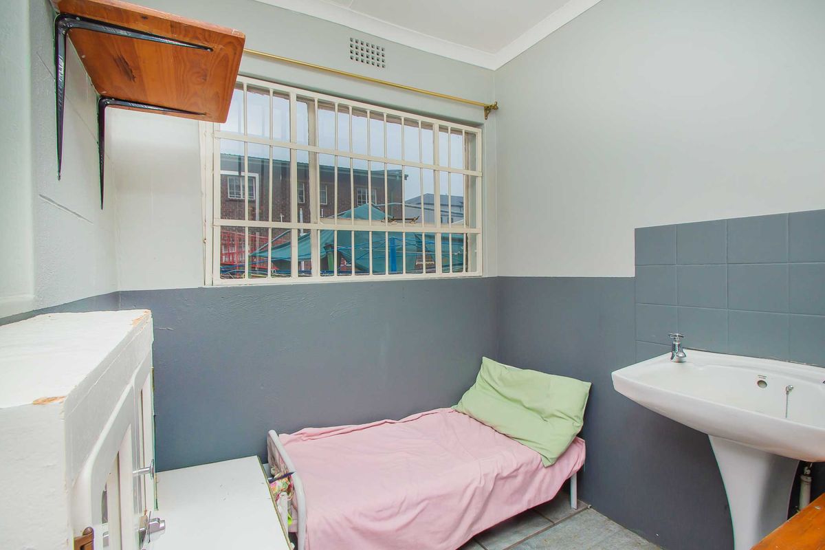 A compact bedroom featuring a pedestal washbasin, security bars on the window, and tiled flooring, requiring cosmetic updates to improve its overall condition.