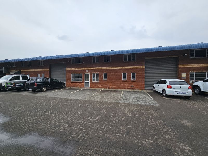 Front elevation of an industrial warehouse unit featuring brick construction, roller shutter doors, and paved parking area.