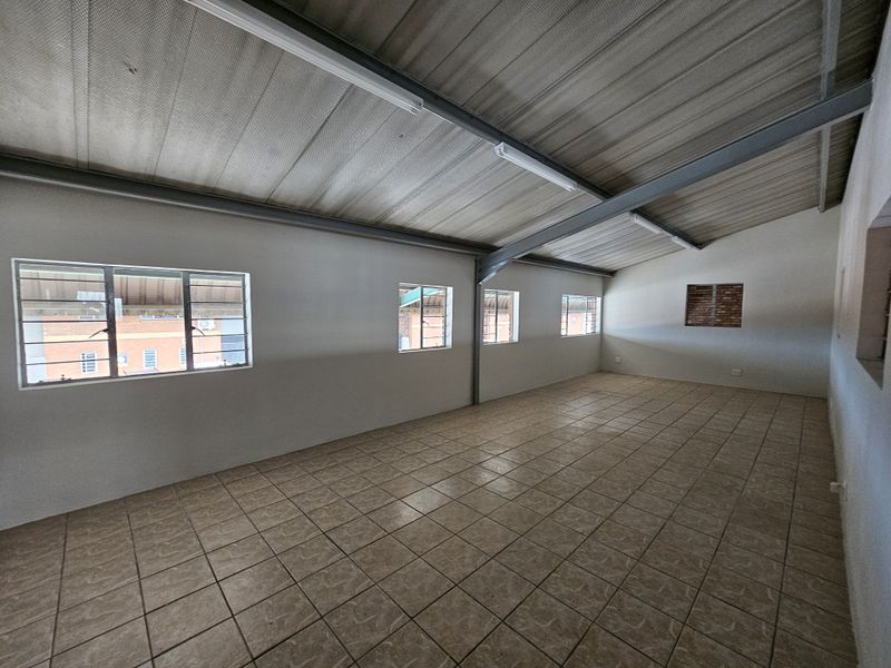 A spacious, empty industrial utility area featuring exposed steel roof trusses, tiled flooring, and multiple steel-framed windows providing natural light.