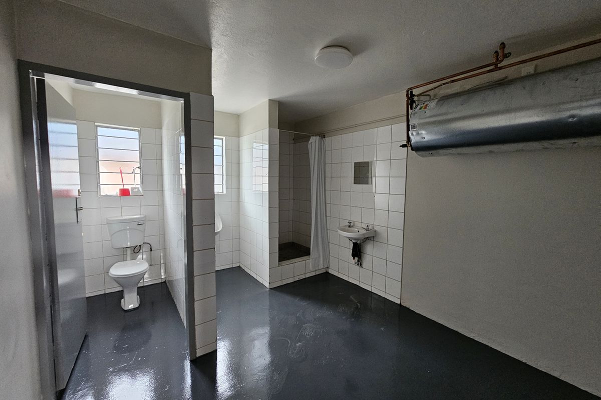 Industrial-style communal bathroom featuring a toilet, urinal, shower cubicle, and wall-mounted basin with exposed water heater plumbing.