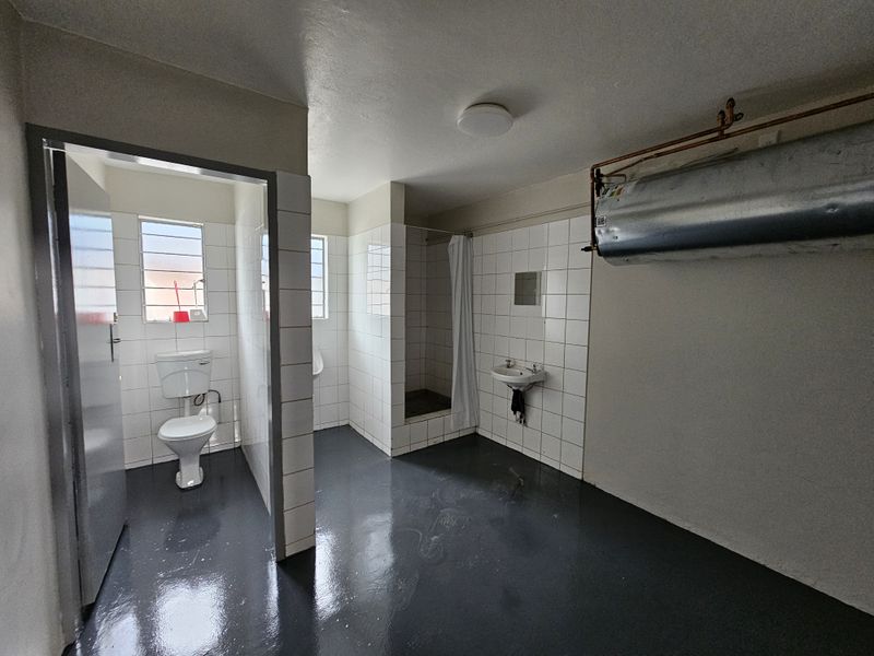 Industrial-style communal bathroom featuring a toilet, urinal, shower cubicle, and wall-mounted basin with exposed water heater plumbing.