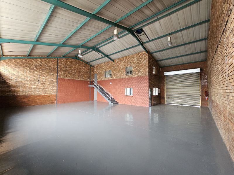 Spacious industrial warehouse unit featuring exposed brick walls, painted concrete flooring, high-clearance steel roof trusses, and a large roller shutter door for easy access.