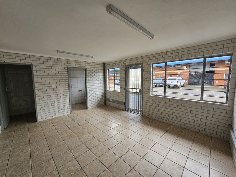 An empty, functional office space featuring tiled flooring, exposed brick walls, fluorescent lighting, and secure window bars overlooking the industrial exterior.