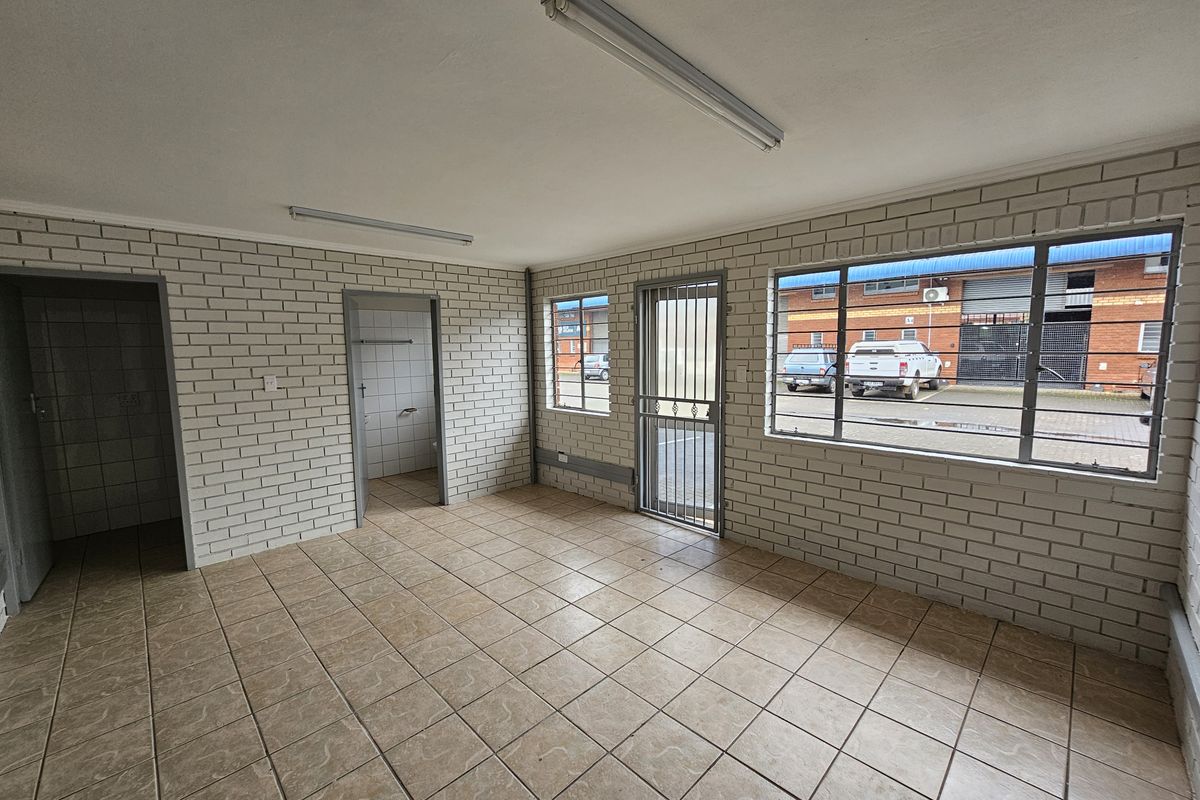 An empty, functional office space featuring tiled flooring, exposed brick walls, fluorescent lighting, and secure window bars overlooking the industrial exterior.