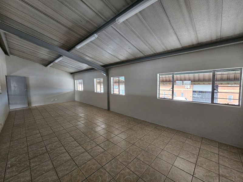 A spacious, empty industrial office area featuring tiled flooring, exposed steel roof beams, and multiple windows providing natural light.