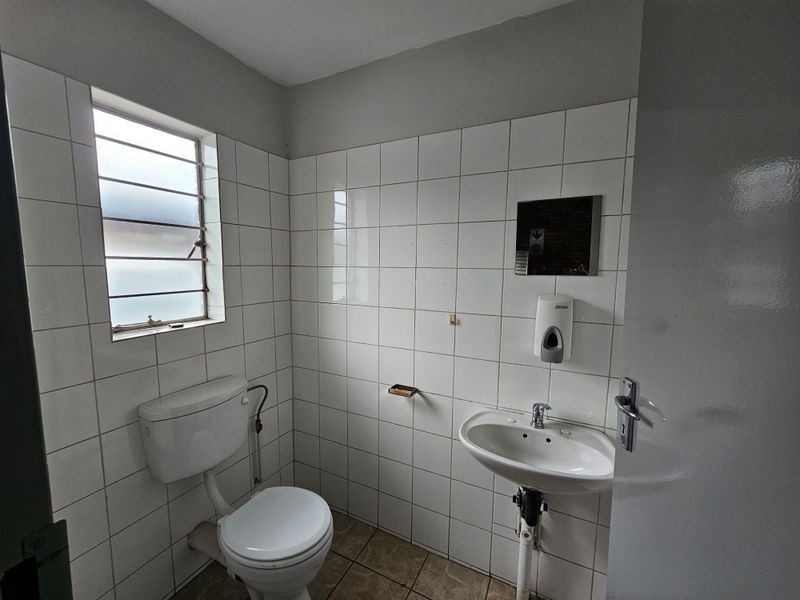 A functional industrial-style bathroom featuring white wall tiles, a pedestal basin, and a standard flush toilet with security bars on the window.