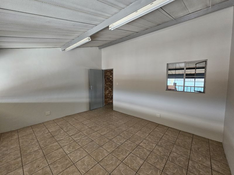 An empty industrial-style office space featuring tiled flooring, exposed metal ceiling beams, fluorescent lighting, and a louvered window.