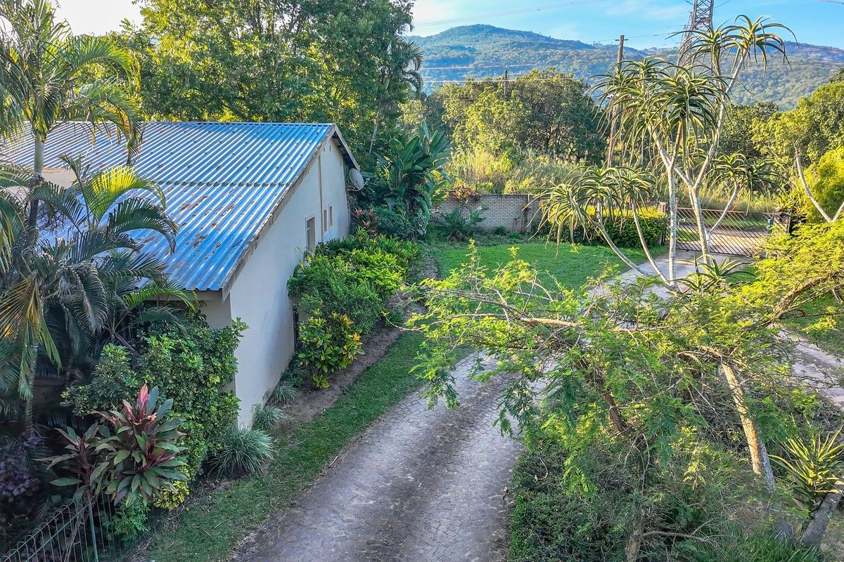 Aerial view of the property showing the main building with a corrugated metal roof, paved driveway, and surrounding garden with mountain views.