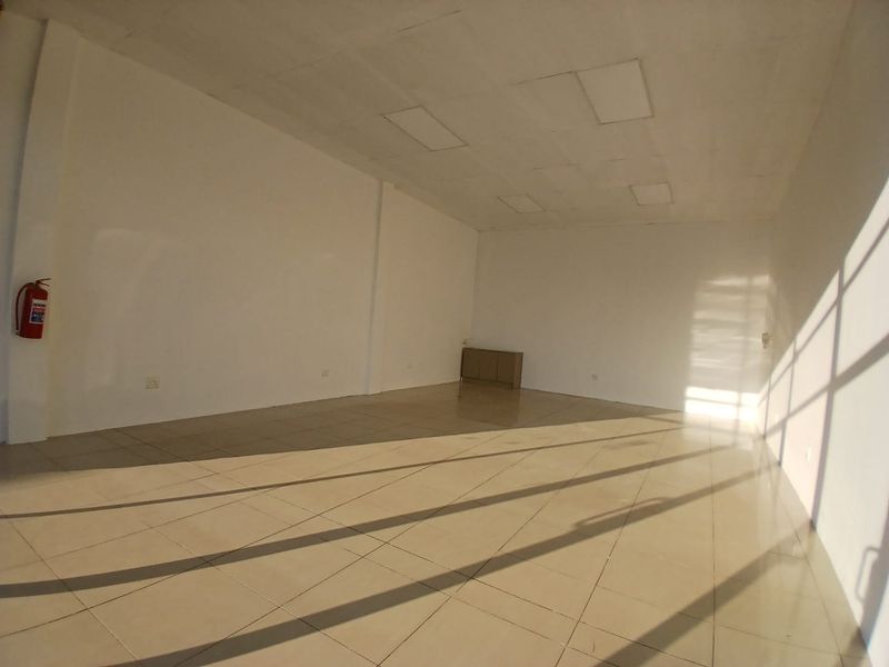 A spacious, empty open-plan retail unit featuring tiled flooring, recessed ceiling lights, and a wall-mounted fire extinguisher.