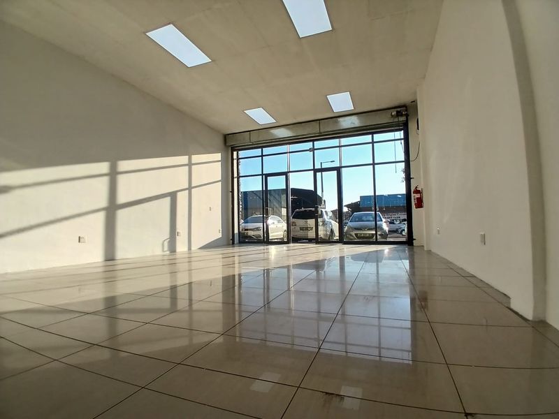 A bright, vacant retail space featuring polished tiled flooring, recessed ceiling lights, and a large glass storefront facade with a roller shutter door.