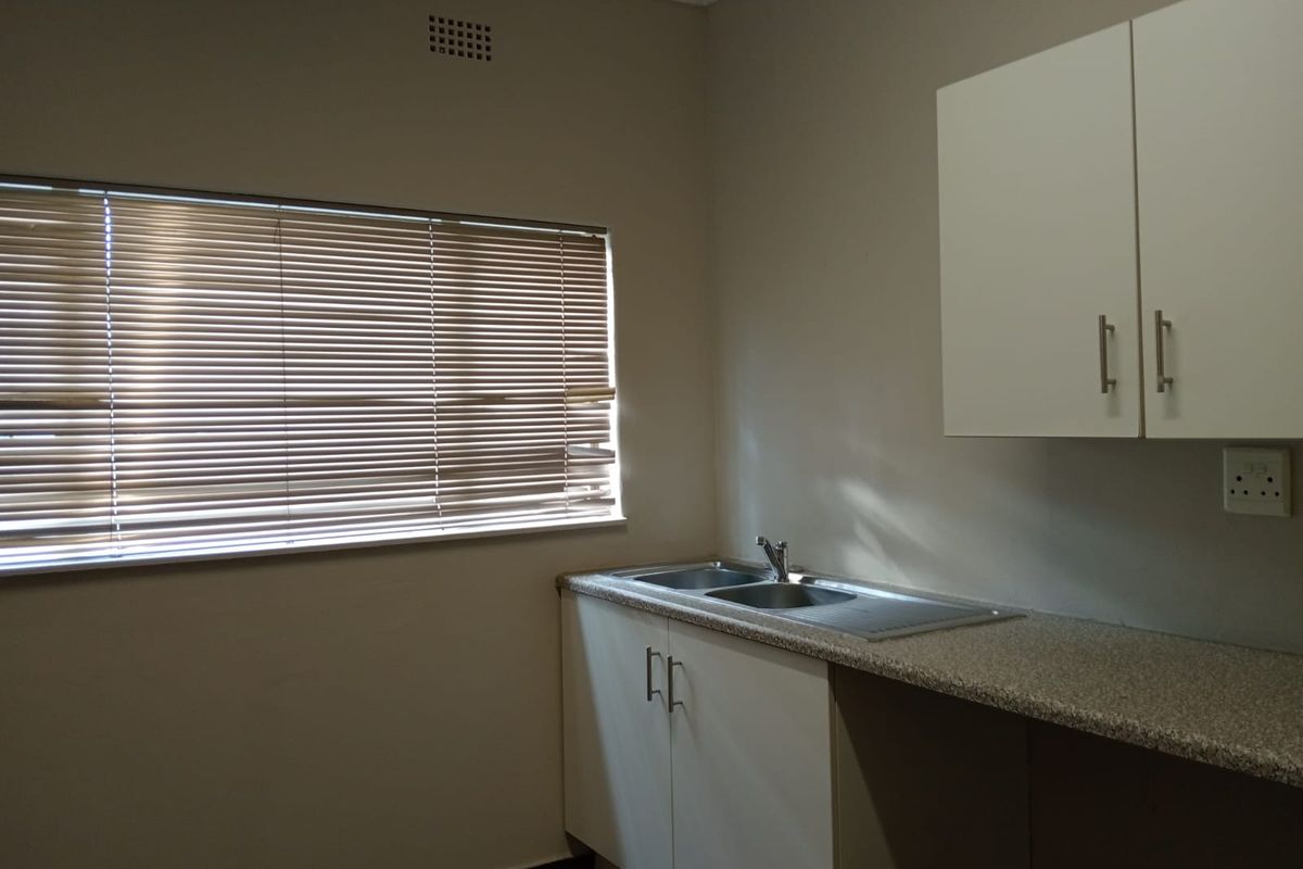 A functional office kitchenette featuring built-in cabinetry, a stainless steel sink, laminate countertops, and natural light through venetian blinds.