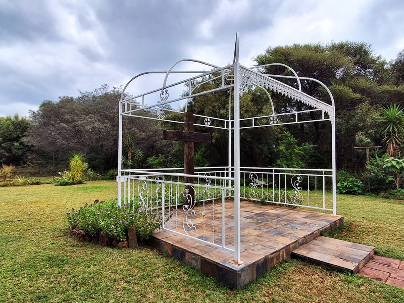 A landscaped garden area featuring a raised tiled platform with a decorative wrought iron gazebo structure and a central wooden cross.