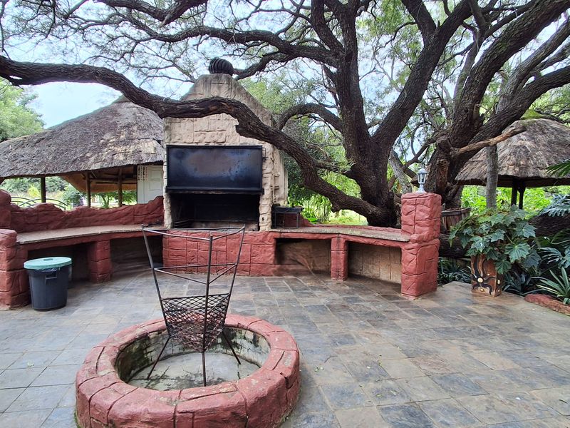 Outdoor entertainment area featuring a built-in braai, stone-clad masonry, fire pit, and thatched roof structure set within a natural garden environment.