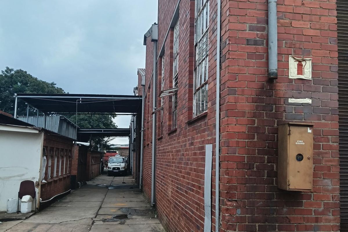 Exterior side access path of an industrial property featuring exposed brick walls, metal window frames, and a wall-mounted fire hose cabinet.