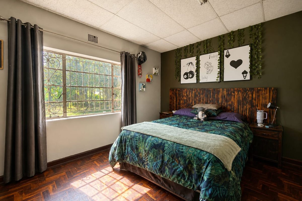 A bedroom featuring parquet flooring, a drop ceiling, and a large window with security burglar bars, offering a functional space with potential for updates.