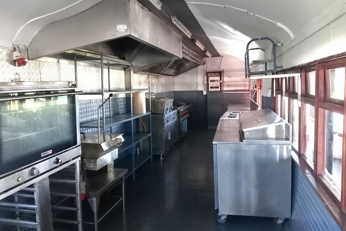 A functional commercial kitchen space featuring stainless steel work surfaces, industrial extractor hoods, and built-in cooking equipment within a narrow, elongated layout.