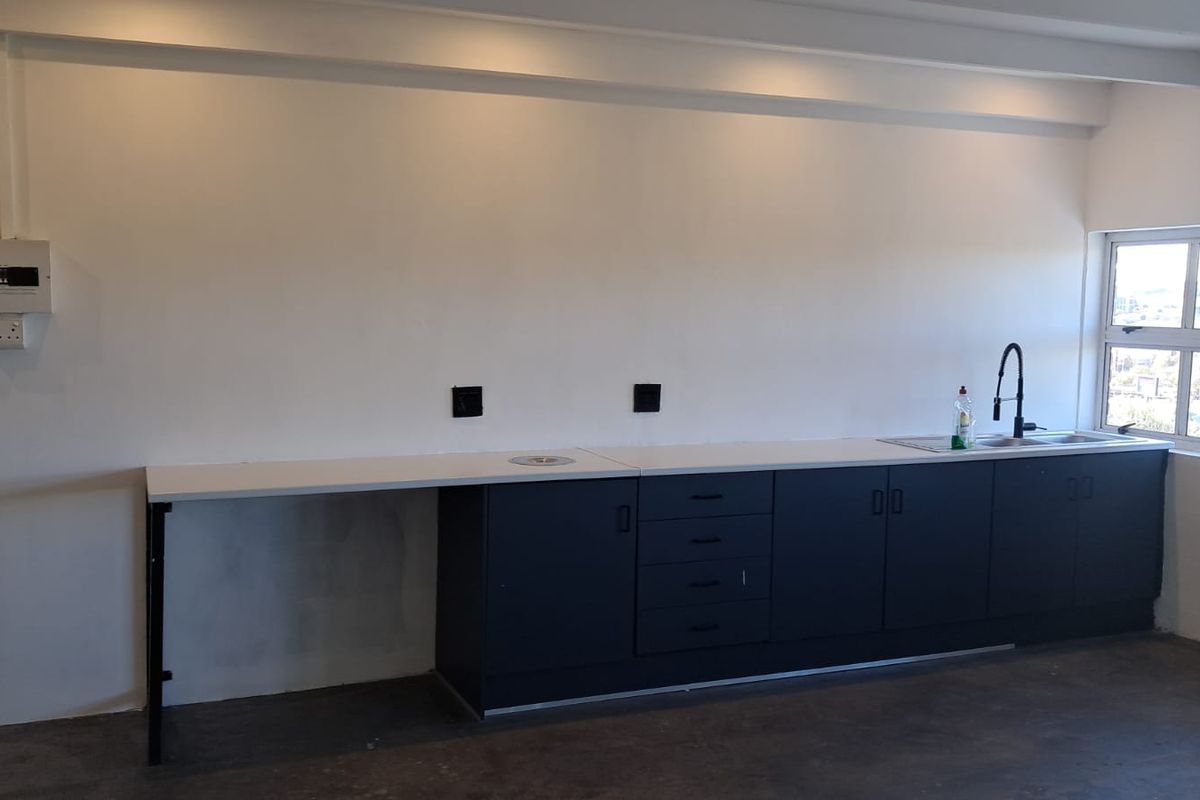A functional kitchenette area featuring dark cabinetry, a white countertop, a stainless steel sink with a modern black faucet, and exposed electrical distribution board.