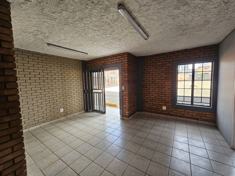 An open-plan industrial office space featuring exposed brick walls, tiled flooring, security gate access, and fluorescent lighting fixtures.