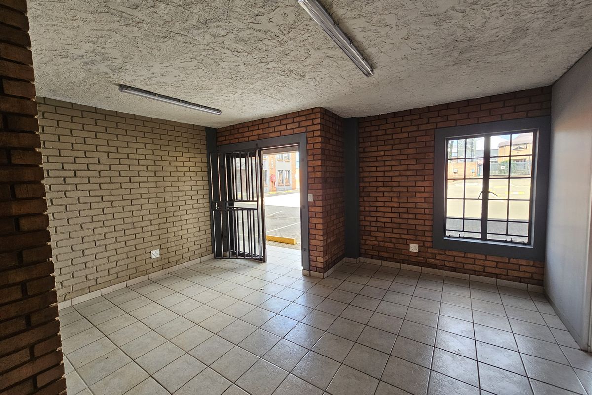 An open-plan industrial office space featuring exposed brick walls, tiled flooring, security gate access, and fluorescent lighting fixtures.
