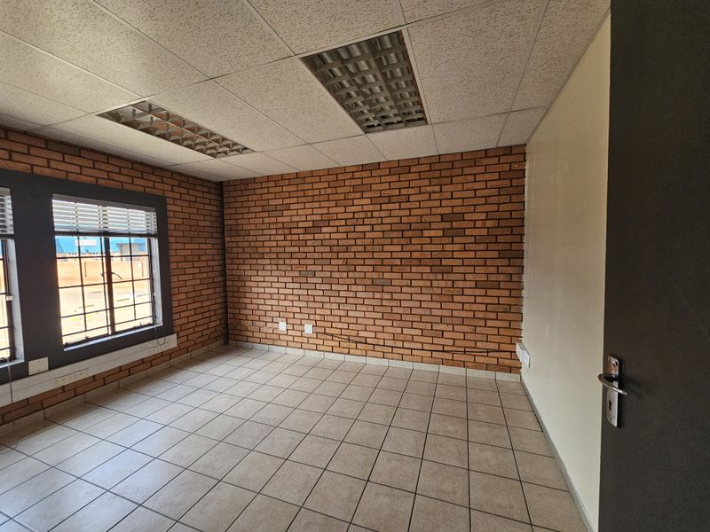 A vacant industrial office space featuring exposed brick walls, tiled flooring, and a suspended ceiling with integrated fluorescent lighting.