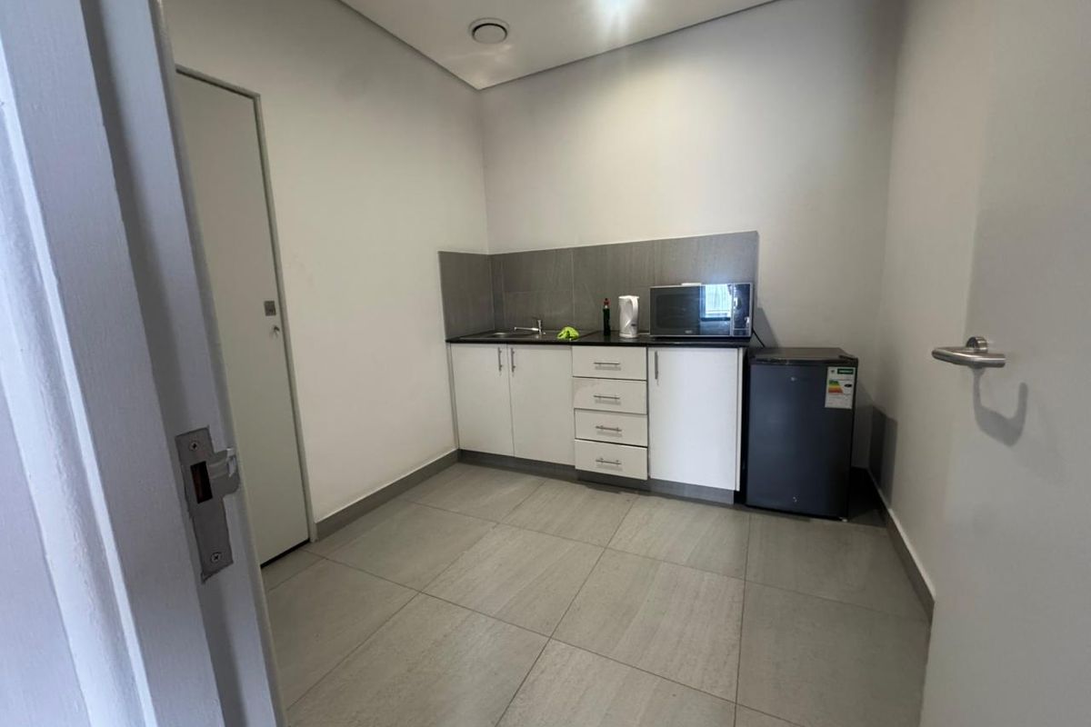 A compact kitchenette featuring modern white cabinetry, dark countertops, tiled splashback, and tiled flooring in a clean, well-maintained industrial space.