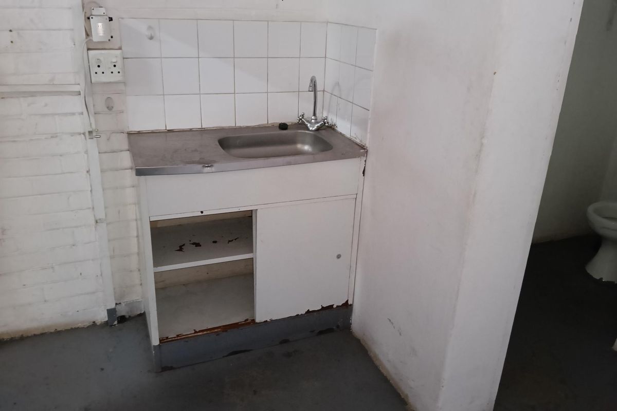 A basic utility kitchenette area featuring a stainless steel sink, tiled splashback, and a damaged base cabinet in an industrial setting.