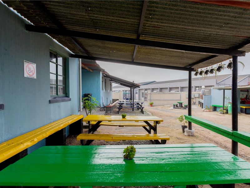 Outdoor covered seating area with corrugated metal roofing, fixed wall-mounted benches, and gravel flooring, suitable for casual restaurant dining.