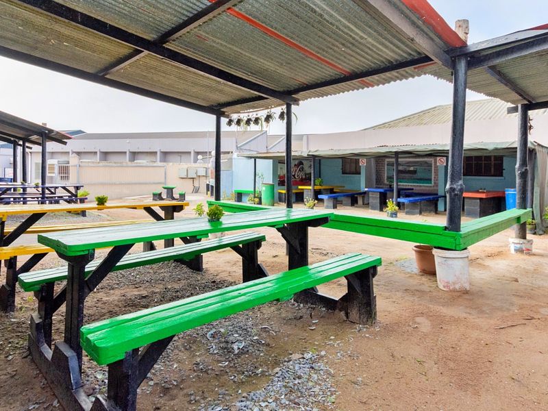 Outdoor covered dining area featuring a corrugated metal roof, steel support structure, and open-air seating space with a dirt and gravel ground surface.
