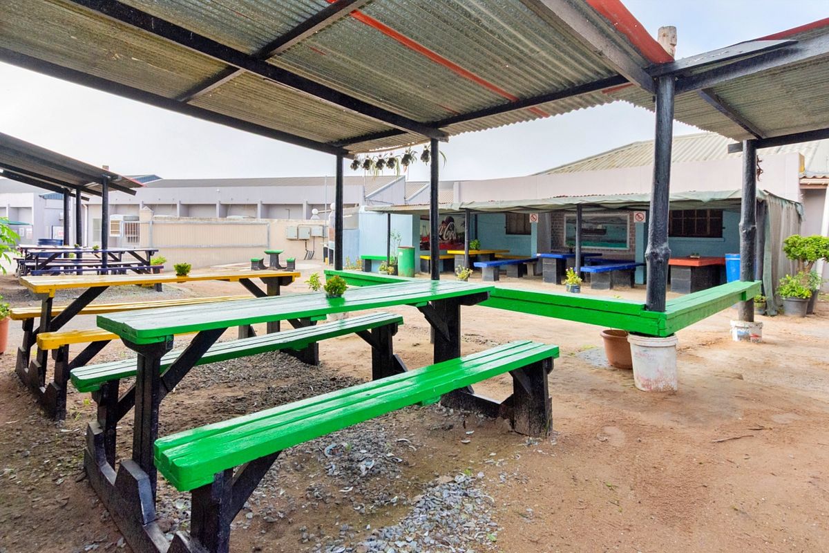 Outdoor covered dining area featuring a corrugated metal roof, steel support structure, and open-air seating space with a dirt and gravel ground surface.