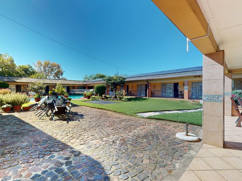 Spacious rear courtyard featuring a cobblestone patio, covered veranda, manicured lawn, and a swimming pool, set within a guest house complex.