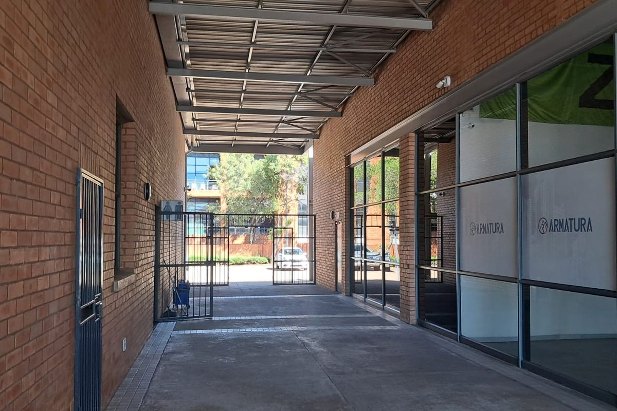 A covered outdoor walkway featuring exposed brick walls, a metal roof structure, and large glass storefront windows, providing access to the industrial unit.