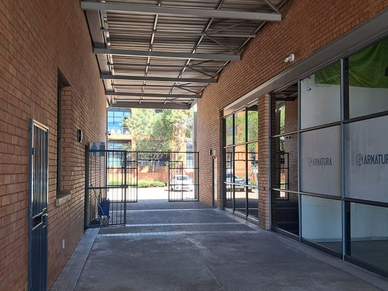 A covered outdoor walkway featuring exposed brick walls, a metal roof structure, and large glass storefront windows, providing access to the industrial unit.