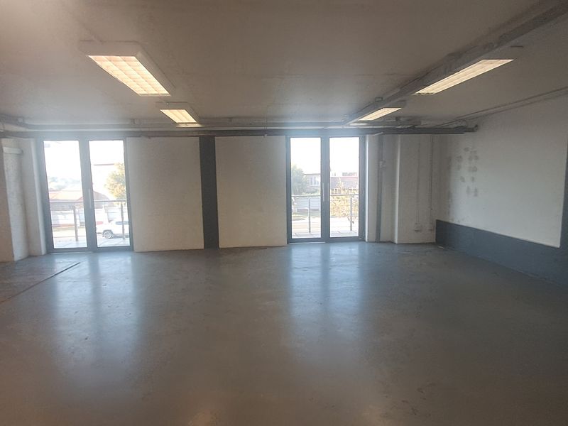 An empty, open-plan industrial space featuring concrete flooring, fluorescent lighting, and double glass doors leading to an exterior area.