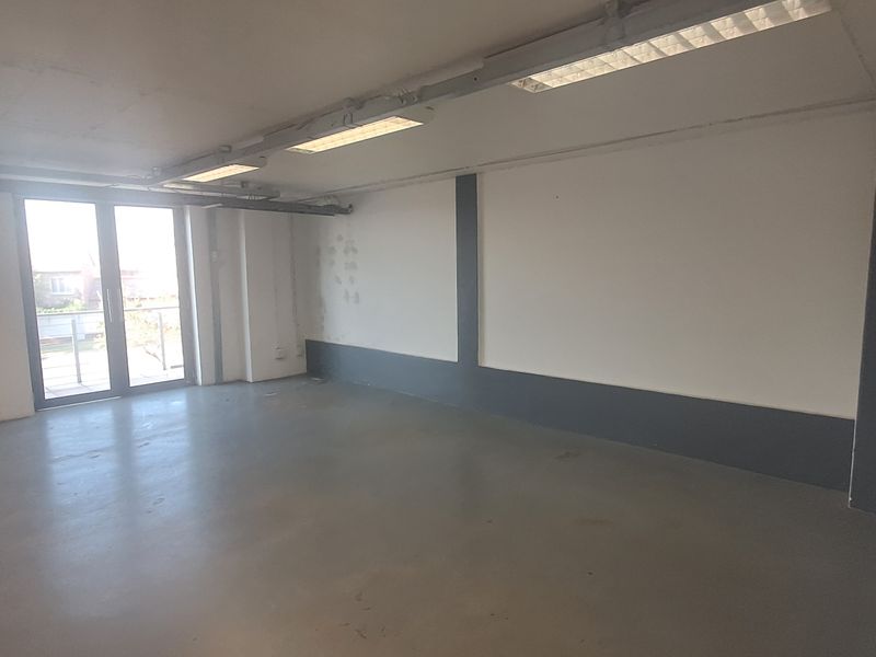 An open-plan industrial office space featuring concrete flooring, fluorescent lighting, and direct access to an outdoor balcony area.