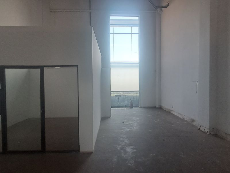 An empty industrial utility space featuring concrete flooring, a partitioned office enclosure, and large windows providing natural light.