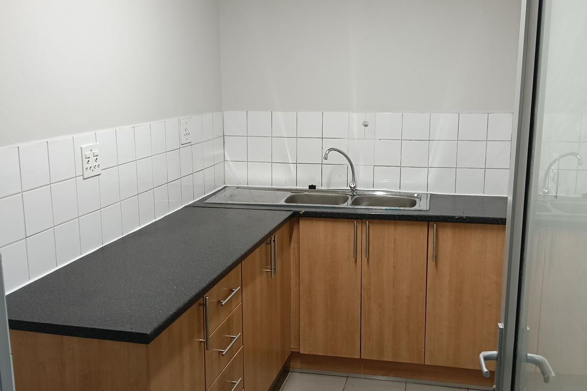 A functional kitchenette featuring wood-finish cabinetry, dark laminate countertops, a stainless steel double sink, and tiled splashbacks in an industrial office setting.