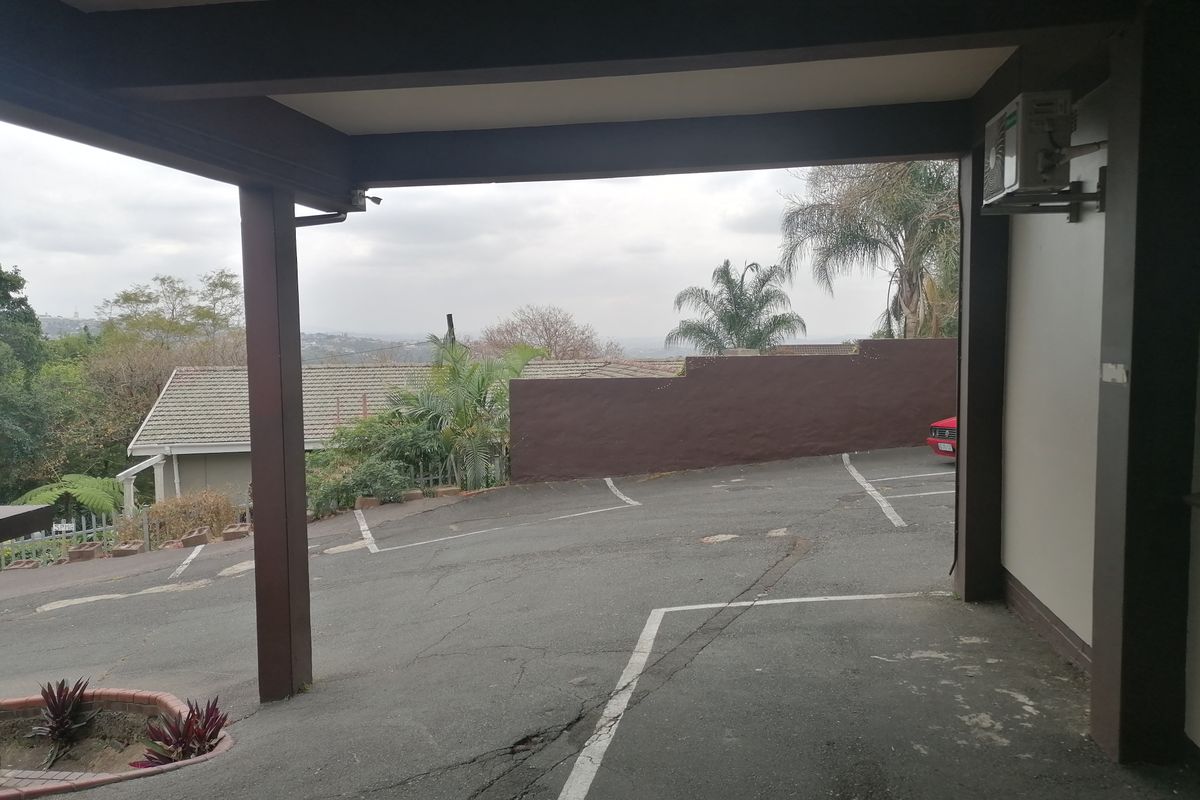 A covered parking area with an asphalt driveway, retaining wall, and exterior air conditioning unit, overlooking a sloped outdoor space.