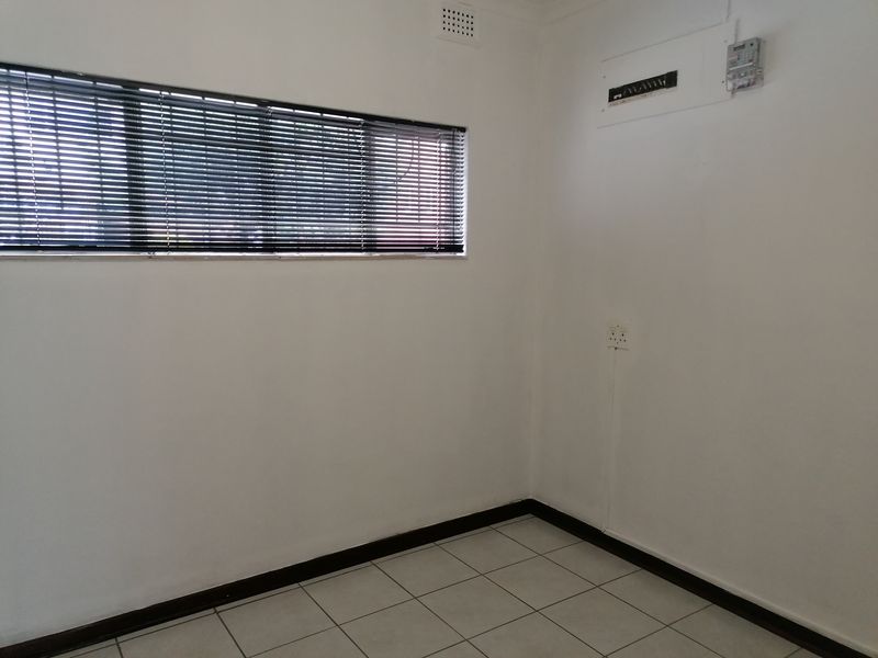 A clean, empty office space featuring white walls, tiled flooring, venetian blinds, and a wall-mounted electrical distribution board.