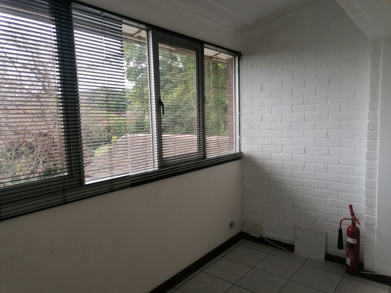A bright, empty office space featuring large windows with venetian blinds, white painted brick walls, and neutral tiled flooring.